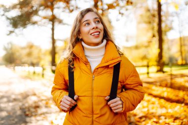 Parlak ceketli şirin bir kadın sarı sonbahar parkında yürüyor. Sırt çantalı genç bir turist güneşli bir günün ve sonbaharın tadını çıkarıyor. Doğa, eğlence konsepti.