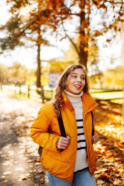Parlak ceketli şirin bir kadın sarı sonbahar parkında yürüyor. Sırt çantalı genç bir turist güneşli bir günün ve sonbaharın tadını çıkarıyor. Doğa, eğlence konsepti.