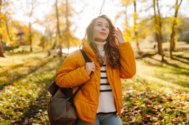 Parlak ceketli şirin bir kadın sarı sonbahar parkında yürüyor. Sırt çantalı genç bir turist güneşli bir günün ve sonbaharın tadını çıkarıyor. Doğa, eğlence konsepti.