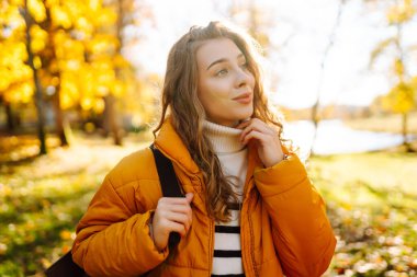 Parlak ceketli şirin bir kadın sarı sonbahar parkında yürüyor. Sırt çantalı genç bir turist güneşli bir günün ve sonbaharın tadını çıkarıyor. Doğa, eğlence konsepti.