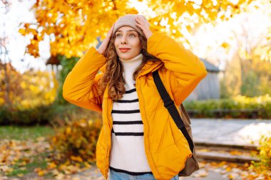 Parlak ceketli şirin bir kadın sarı sonbahar parkında yürüyor. Sırt çantalı genç bir turist güneşli bir günün ve sonbaharın tadını çıkarıyor. Doğa, eğlence konsepti.