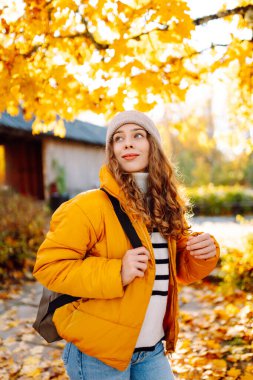 Parlak ceketli şirin bir kadın sarı sonbahar parkında yürüyor. Sırt çantalı genç bir turist güneşli bir günün ve sonbaharın tadını çıkarıyor. Doğa, eğlence konsepti.