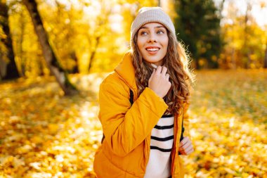 Parlak ceketli şirin bir kadın sarı sonbahar parkında yürüyor. Sırt çantalı genç bir turist güneşli bir günün ve sonbaharın tadını çıkarıyor. Doğa, eğlence konsepti.