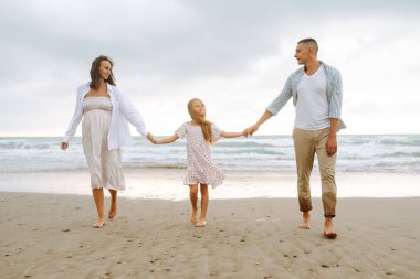 Kumsalda el ele tutuşarak yürüyen mutlu bir aile. Hamile karısı ve kızıyla birlikte dışarıda özgürce vakit geçiren bir adam. Aile, hafta sonu konsepti.