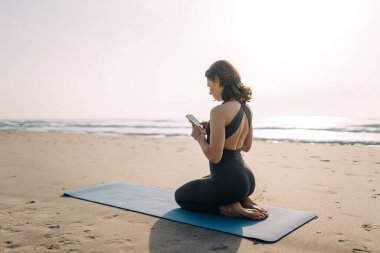 Spor kıyafetli rahat bir kadın gün batımında yoga minderinde yoga pozu veriyor. Güzel bir kadın spor yapıyor, kumsalda egzersiz yapıyor. Spor, rahatlama ve doğa konsepti.