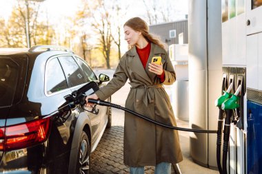Benzin istasyonunda telefonu elinde tutan genç bir kadın. Benzin dolduran mobil uygulama kullanan neşeli bir kadın. Teknoloji kavramı, ulaşım.