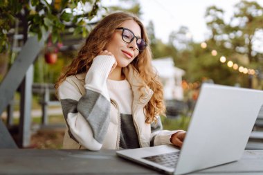 Bilgisayarlı genç bir kadın parkta bir masada oturmuş, doğanın tadını çıkarıyor. Dışarıda dizüstü bilgisayarda çalışan serbest çalışan bir kadın. Uzak çalışma ve teknoloji kavramı.