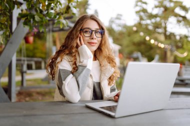 Bilgisayarlı genç bir kadın parkta bir masada oturmuş, doğanın tadını çıkarıyor. Dışarıda dizüstü bilgisayarda çalışan serbest çalışan bir kadın. Uzak çalışma ve teknoloji kavramı.