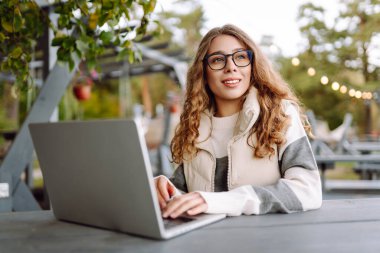 Bilgisayarlı genç bir kadın parkta bir masada oturmuş, doğanın tadını çıkarıyor. Dışarıda dizüstü bilgisayarda çalışan serbest çalışan bir kadın. Uzak çalışma ve teknoloji kavramı.