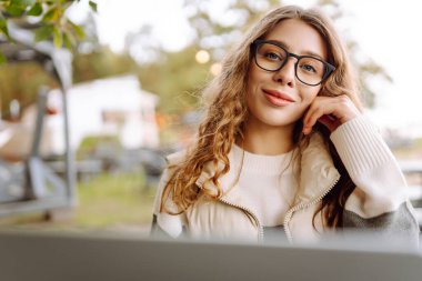 Bilgisayarlı genç bir kadın parkta bir masada oturmuş, doğanın tadını çıkarıyor. Dışarıda dizüstü bilgisayarda çalışan serbest çalışan bir kadın. Uzak çalışma ve teknoloji kavramı.