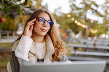 Bilgisayarlı genç bir kadın parkta bir masada oturmuş, doğanın tadını çıkarıyor. Dışarıda dizüstü bilgisayarda çalışan serbest çalışan bir kadın. Uzak çalışma ve teknoloji kavramı.
