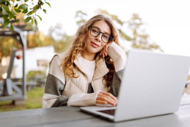 Bilgisayarlı genç bir kadın parkta bir masada oturmuş, doğanın tadını çıkarıyor. Dışarıda dizüstü bilgisayarda çalışan serbest çalışan bir kadın. Uzak çalışma ve teknoloji kavramı.