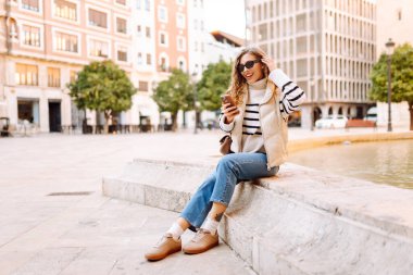 Güneş gözlüklü şık bir kadın ve telefonla güneşli Avrupa caddelerinde yürüyüş yapıyor. Genç turist blogları ya da açık havada mesajlaşmalar. Moda, tarz ve turizm konsepti.