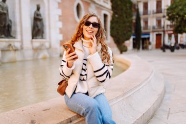 Güneş gözlüklü şık bir kadın ve telefonla güneşli Avrupa caddelerinde yürüyüş yapıyor. Genç turist blogları ya da açık havada mesajlaşmalar. Moda, tarz ve turizm konsepti.