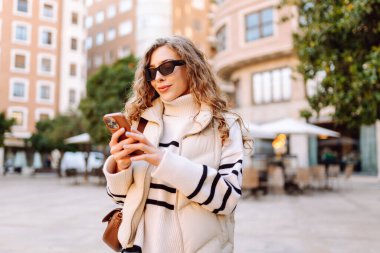 Güneş gözlüklü şık bir kadın ve telefonla güneşli Avrupa caddelerinde yürüyüş yapıyor. Genç turist blogları ya da açık havada mesajlaşmalar. Moda, tarz ve turizm konsepti.
