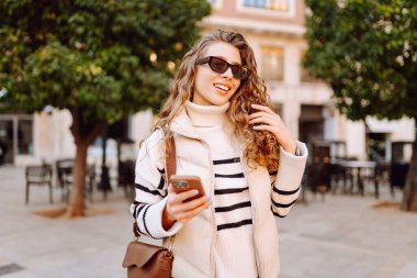 Güneş gözlüklü şık bir kadın ve telefonla güneşli Avrupa caddelerinde yürüyüş yapıyor. Genç turist blogları ya da açık havada mesajlaşmalar. Moda, tarz ve turizm konsepti.