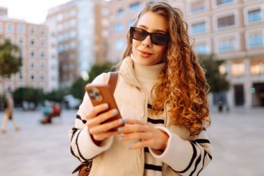Güneş gözlüklü şık bir kadın ve telefonla güneşli Avrupa caddelerinde yürüyüş yapıyor. Genç turist blogları ya da açık havada mesajlaşmalar. Moda, tarz ve turizm konsepti.