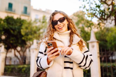 Güneş gözlüklü şık bir kadın ve telefonla güneşli Avrupa caddelerinde yürüyüş yapıyor. Genç turist blogları ya da açık havada mesajlaşmalar. Moda, tarz ve turizm konsepti.