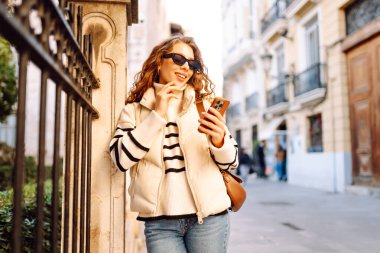 Güneş gözlüklü şık bir kadın ve telefonla güneşli Avrupa caddelerinde yürüyüş yapıyor. Genç turist blogları ya da açık havada mesajlaşmalar. Moda, tarz ve turizm konsepti.
