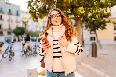 Güneş gözlüklü şık bir kadın ve telefonla güneşli Avrupa caddelerinde yürüyüş yapıyor. Genç turist blogları ya da açık havada mesajlaşmalar. Moda, tarz ve turizm konsepti.