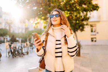 Güneş gözlüklü şık bir kadın ve telefonla güneşli Avrupa caddelerinde yürüyüş yapıyor. Genç turist blogları ya da açık havada mesajlaşmalar. Moda, tarz ve turizm konsepti.