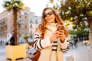 Güneş gözlüklü şık bir kadın ve telefonla güneşli Avrupa caddelerinde yürüyüş yapıyor. Genç turist blogları ya da açık havada mesajlaşmalar. Moda, tarz ve turizm konsepti.
