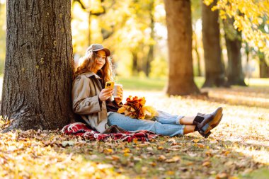 Sarı yapraklarla çevrili bir sonbahar ormanında, bir battaniyenin üzerinde oturan şapkalı genç bir kadın. Bir fincan sıcak içecekle neşeli bir kadın ve açık havada güneşin tadını çıkaran bir telefon. Yaşam biçimi.