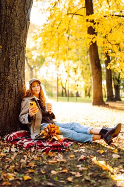 Sarı yapraklarla çevrili bir sonbahar ormanında, bir battaniyenin üzerinde oturan şapkalı genç bir kadın. Bir fincan sıcak içecekle neşeli bir kadın ve açık havada güneşin tadını çıkaran bir telefon. Yaşam biçimi.