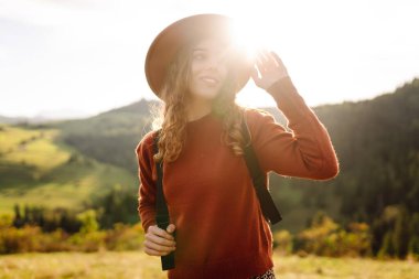 Yeşil tepelerde yürüyüp gün batımının tadını çıkaran sırt çantalı ve şapkalı genç bayan gezginin arka görüntüsü. Güzel kadın dağ manzarasına karşı özgürlüğü hissediyor. Yürüyüş ve seyahat konsepti.