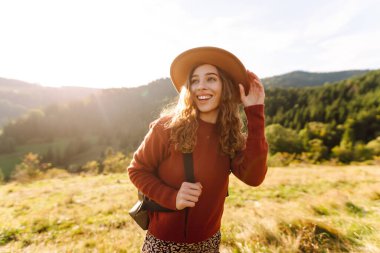 Yeşil tepelerde yürüyüp gün batımının tadını çıkaran sırt çantalı ve şapkalı genç bayan gezginin arka görüntüsü. Güzel kadın dağ manzarasına karşı özgürlüğü hissediyor. Yürüyüş ve seyahat konsepti.