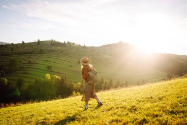 Yeşil tepelerde yürüyüp gün batımının tadını çıkaran sırt çantalı ve şapkalı genç bayan gezginin arka görüntüsü. Güzel kadın dağ manzarasına karşı özgürlüğü hissediyor. Yürüyüş ve seyahat konsepti.