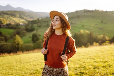 Yeşil tepelerde yürüyüp gün batımının tadını çıkaran sırt çantalı ve şapkalı genç bayan gezginin arka görüntüsü. Güzel kadın dağ manzarasına karşı özgürlüğü hissediyor. Yürüyüş ve seyahat konsepti.