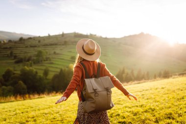 Yeşil tepelerde yürüyüp gün batımının tadını çıkaran sırt çantalı ve şapkalı genç bayan gezginin arka görüntüsü. Güzel kadın dağ manzarasına karşı özgürlüğü hissediyor. Yürüyüş ve seyahat konsepti.