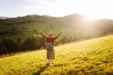 Yeşil tepelerde yürüyüp gün batımının tadını çıkaran sırt çantalı ve şapkalı genç bayan gezginin arka görüntüsü. Güzel kadın dağ manzarasına karşı özgürlüğü hissediyor. Yürüyüş ve seyahat konsepti.