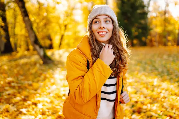 Parlak ceketli şirin bir kadın sarı sonbahar parkında yürüyor. Sırt çantalı genç bir turist güneşli bir günün ve sonbaharın tadını çıkarıyor. Doğa, eğlence konsepti.