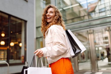 Şık giyinmiş neşeli bir kadın alışveriş merkezinin arka planında alışveriş torbaları tutuyor. Güzel bir kadın alışveriş yaptıktan sonra eğleniyor ve güneşli havanın tadını çıkarıyor. Alışveriş zamanı. Tüketim kavramı.