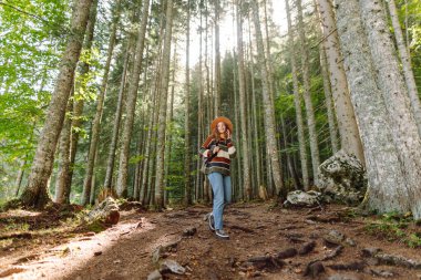 Sırt çantası ve şapkası olan genç bir kadın sık bir ormanda kayalık bir yolda yürüyor. Dişi bir gezgin doğa ve yürüyüş yapmaktan zevk alır. Seyahat, yürüyüş ve doğa konsepti. Etkin yaşam biçimi.