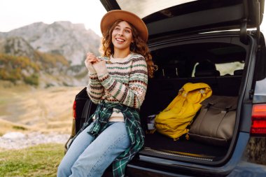Yolculukta olan tatlı bir kadın bir arabanın bagajında oturur ve dağ manzarasının tadını çıkarır. Sırt çantalı bir bayan gezgin kendini özgür hisseder ve bir arabanın bagajında eğlenir. Etkin yaşam biçimi.