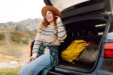 Yolculukta olan tatlı bir kadın bir arabanın bagajında oturur ve dağ manzarasının tadını çıkarır. Sırt çantalı bir bayan gezgin kendini özgür hisseder ve bir arabanın bagajında eğlenir. Etkin yaşam biçimi.