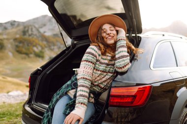 Yolculukta olan tatlı bir kadın bir arabanın bagajında oturur ve dağ manzarasının tadını çıkarır. Sırt çantalı bir bayan gezgin kendini özgür hisseder ve bir arabanın bagajında eğlenir. Etkin yaşam biçimi.