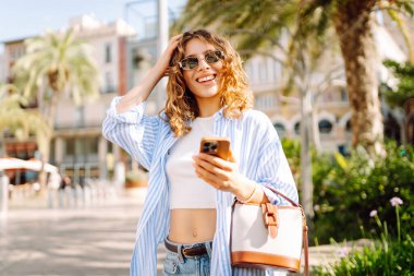 Gündelik kıyafetlerle güzel bir kadın telefonuyla şehrin güneşli sokaklarında eğleniyor. Genç bir turist gün batımının ve telefonuyla mesajlaşmanın tadını çıkarıyor. Moda ve güzellik konsepti. Yaşam biçimi.