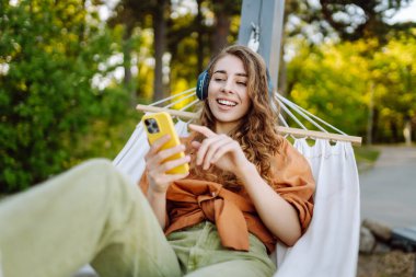 Telefonlu ve kulaklıklı gülümseyen kadın hamakta uzanıp güneşli parkta hafta sonunun tadını çıkarıyor. Hamakta müzik dinleyen ya da podcast yapan güzel bir kadın. Teknoloji konsepti, hafta sonu.