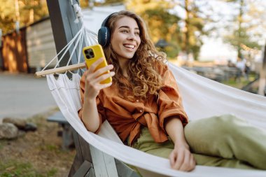 Telefonlu ve kulaklıklı gülümseyen kadın hamakta uzanıp güneşli parkta hafta sonunun tadını çıkarıyor. Hamakta müzik dinleyen ya da podcast yapan güzel bir kadın. Teknoloji konsepti, hafta sonu.