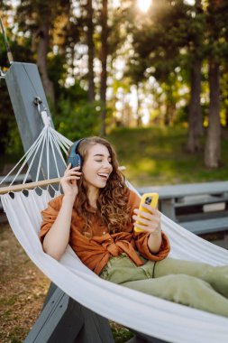 Telefonlu ve kulaklıklı gülümseyen kadın hamakta uzanıp güneşli parkta hafta sonunun tadını çıkarıyor. Hamakta müzik dinleyen ya da podcast yapan güzel bir kadın. Teknoloji konsepti, hafta sonu.