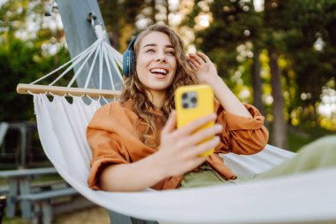 Telefonlu ve kulaklıklı gülümseyen kadın hamakta uzanıp güneşli parkta hafta sonunun tadını çıkarıyor. Hamakta müzik dinleyen ya da podcast yapan güzel bir kadın. Teknoloji konsepti, hafta sonu.
