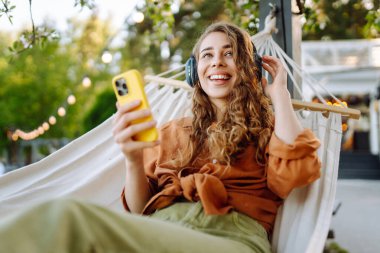 Telefonlu ve kulaklıklı gülümseyen kadın hamakta uzanıp güneşli parkta hafta sonunun tadını çıkarıyor. Hamakta müzik dinleyen ya da podcast yapan güzel bir kadın. Teknoloji konsepti, hafta sonu.