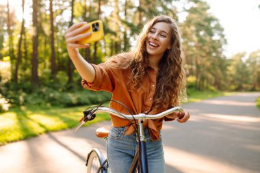 Telefonlu ve bisikletli neşeli kadın güneşli parkta selfie çekiyor. Bisiklete binen ve dışarıda havanın tadını çıkaran genç bir kadın. Yürüme konsepti, selfie. Etkin yaşam biçimi.