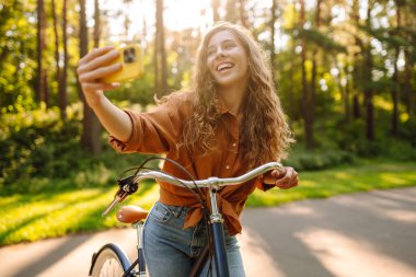 Telefonlu ve bisikletli neşeli kadın güneşli parkta selfie çekiyor. Bisiklete binen ve dışarıda havanın tadını çıkaran genç bir kadın. Yürüme konsepti, selfie. Etkin yaşam biçimi.