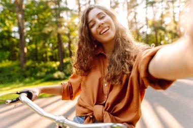 Telefonlu ve bisikletli neşeli kadın güneşli parkta selfie çekiyor. Bisiklete binen ve dışarıda havanın tadını çıkaran genç bir kadın. Yürüme konsepti, selfie. Etkin yaşam biçimi.
