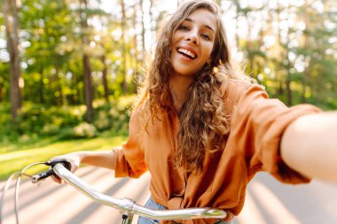 Telefonlu ve bisikletli neşeli kadın güneşli parkta selfie çekiyor. Bisiklete binen ve dışarıda havanın tadını çıkaran genç bir kadın. Yürüme konsepti, selfie. Etkin yaşam biçimi.